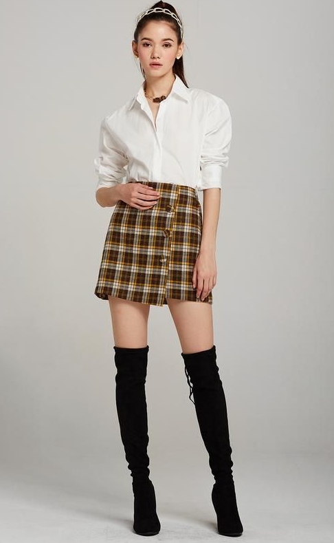 A model standing in white room wearing a shirt, mini skirt and thigh-high boots