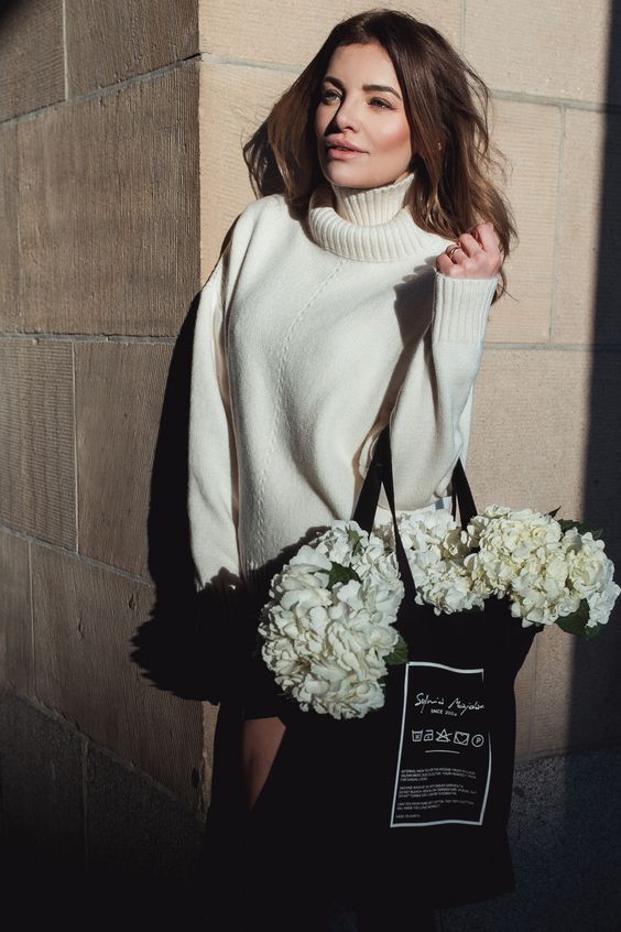 A girl standing beside a wall, wearing a turtle neck sweater and hanging a shoping bag with flowers in left hand.